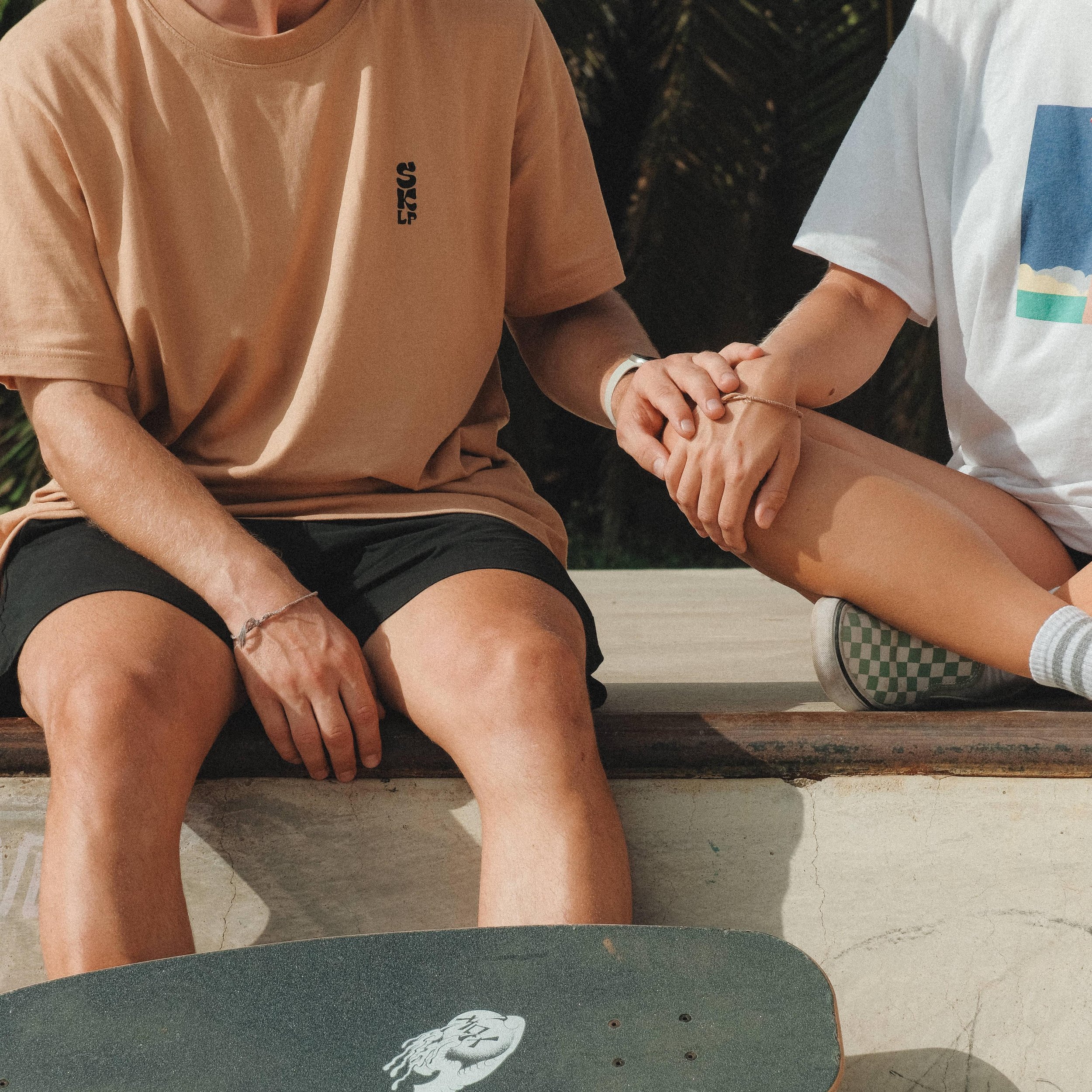 a couple holding hands at the skatepark, sitting at the edge of a bowl and wearing the surfskate las palmas merch tshirt.