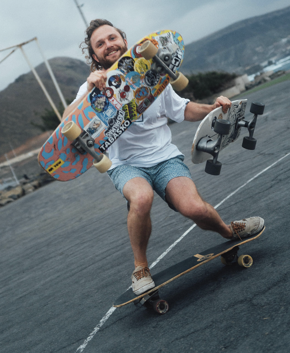 A guy skating with two surfskates in his hand.